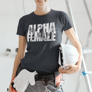 May include: Dark gray t-shirt with the text "ALPHA FEMALE" in white, featuring a silhouette design. The person is wearing work pants, a tool belt, and holding a white hard hat and gloves. A ladder is in the background.