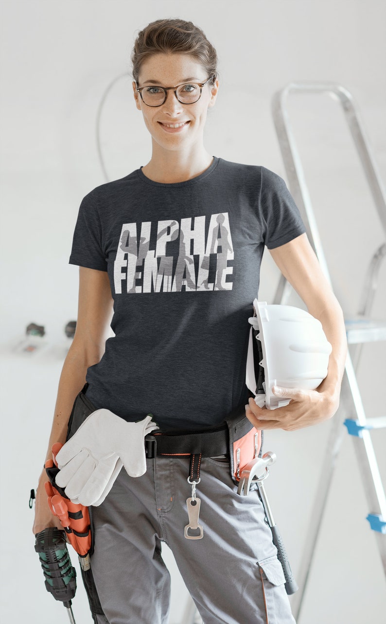 May include: Dark gray t-shirt with the text "ALPHA FEMALE" in white, featuring a silhouette design. The person is wearing work pants, a tool belt, and holding a white hard hat and gloves. A ladder is in the background.