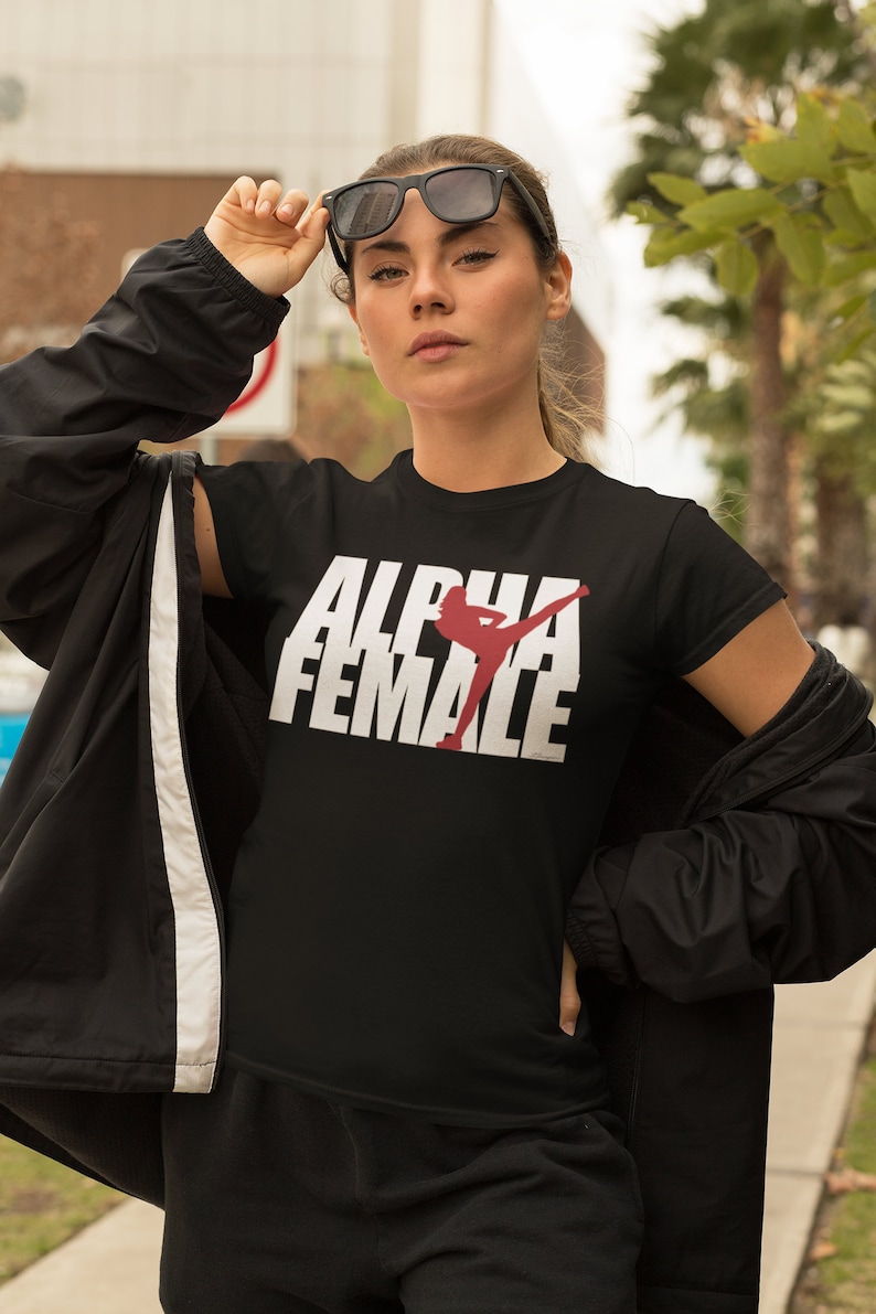 May include: Black t-shirt with the text "ALPHA FEMALE" in white block letters and a red silhouette of a woman kicking. The person is wearing black sunglasses and a black jacket.