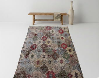May include: A rectangular, vintage-style rug with a geometric pattern in shades of gray, red, beige, and green. The rug is placed on a white floor. A wooden bench and a tall, cream-colored vase are in the background.