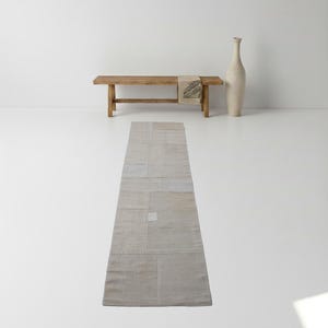 May include: A neutral-toned, rectangular patchwork rug with a light beige color scheme. The rug is made of various rectangular fabric pieces sewn together. A wooden bench with a patterned throw and a tall, cream-colored vase are in the background.