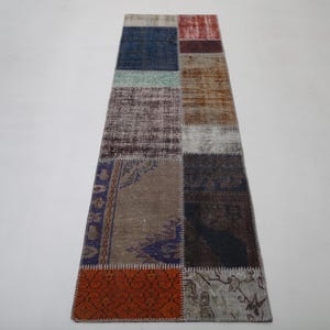 Può includere: Un tappeto rettangolare patchwork con una varietà di colori e motivi. Il tappeto presenta quadrati blu, rossi, marroni e beige. Il tappeto è probabilmente realizzato in lana o materiale simile.