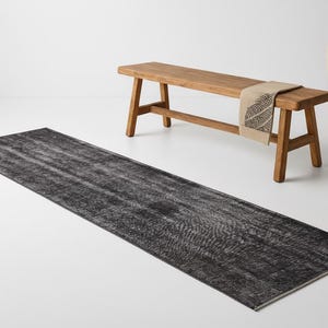 May include: A long, rectangular, distressed black rug with a faded pattern. A wooden bench with a beige patterned runner sits behind the rug. A tall, cream-colored vase is also in the background.