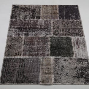 Puede incluir: Una alfombra de retazos con un aspecto envejecido y vintage. Está compuesta por varios parches rectangulares en tonos grises, marrones y negros, cosidos con hilo blanco. El diseño presenta patrones y texturas sutiles.