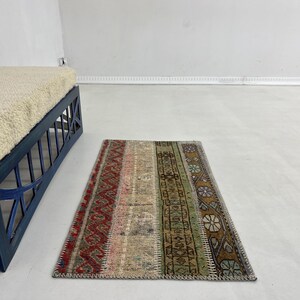 Könnte beinhalten: Ein rechteckiger, mehrfarbiger Teppich mit Patchwork-Design. Der Teppich hat vertikale Streifen in Rot, Rosa, Beige, Grün und Braun. Der Teppich liegt auf einem hellen Boden. Im Hintergrund befindet sich eine blaue Bank mit cremefarbenem Kissen.