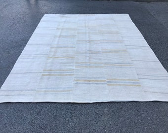 Può includere: Un tappeto bianco tessuto con un sottile motivo a righe in tonalità beige e grigio. Il tappeto ha una forma rettangolare e una superficie morbida e testurizzata.