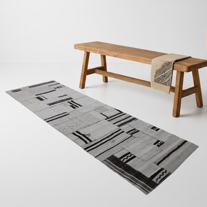 May include: A rectangular gray rug with black geometric patterns is displayed on a white surface. A wooden bench with a beige throw and a tall, cream-colored vase are in the background. The rug's design includes straight and zigzag lines.