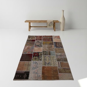 May include: A rectangular patchwork rug with a variety of colors and patterns. The rug is made up of many different pieces of fabric sewn together. The colors include brown, red, blue, and beige. A wooden bench and a tall vase are in the background.