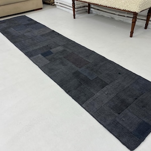 May include: A long, rectangular, dark blue patchwork rug made from denim fabric. The rug is made up of many different sized squares and rectangles of denim, all sewn together. The rug is lying on a white floor.