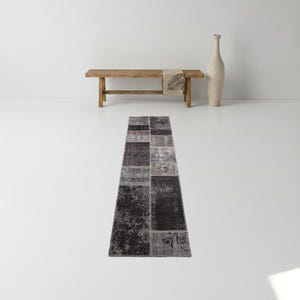 May include: A rectangular patchwork rug with a variety of dark gray, black, and light gray patches. The rug is placed on a white floor. A wooden bench with a beige throw and a tall, cream-colored vase are in the background.