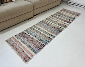 Può includere: Un tappeto lungo e stretto a strisce con frange sui bordi. Il tappeto ha uno sfondo crema con strisce blu, rosse e nere.