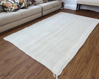 Può includere: Un grande tappeto rettangolare di colore bianco sporco con una sottile trama tessuta. Il tappeto è steso su un pavimento in legno duro.