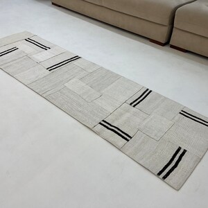 May include: A rectangular, light gray runner rug with a patchwork design. The rug features black stripes across several of the patches. The rug is made of a woven material and is placed on a white surface.