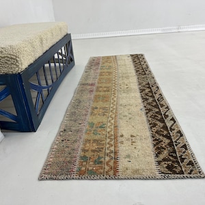 May include: A rectangular, multi-colored rug with a patchwork design. The rug features various shades of beige, brown, and muted colors. The rug is placed on a white floor next to a blue bench with a cream-colored cushion.