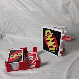 May include: A red and black UNO card game set. The set includes a red card holder with a stack of cards, and a white and black card holder with the word UNO in yellow. The cards have the word UNO and the number 2.