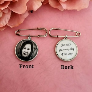 Op de afbeelding: Een zilveren veiligheidsspeld met een ronde hanger eraan. De voorkant van de hanger heeft een zwart-witfoto van een vrouw. De achterkant van de hanger heeft de tekst "Ik ben bij je bij elke stap van de weg."
