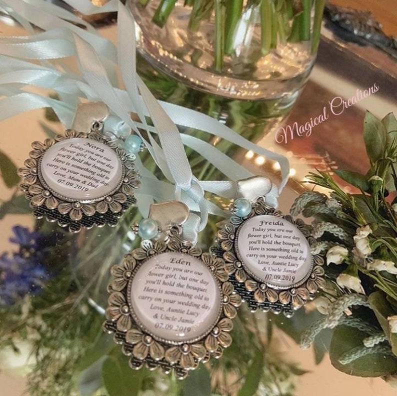 K&ouml;nnte beinhalten: Drei silberne bl&uuml;tenf&ouml;rmige Anh&auml;nger mit blauen B&auml;ndern und wei&szlig;en Glaseins&auml;tzen. Jeder Anh&auml;nger tr&auml;gt eine personalisierte Botschaft f&uuml;r ein Blumenm&auml;dchen, einschlie&szlig;lich des Namens des M&auml;dchens, des Datums der Hochzeit und einer Botschaft des Gebers. Die Anh&auml;nger befinden sich auf einer reflektierenden Oberfl&auml;che.