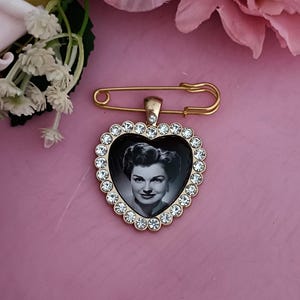 May include: A gold-toned heart-shaped brooch with a black and white portrait of a woman. The heart is framed with clear crystals. A gold safety pin is attached to the top of the brooch. The background is a soft pink.
