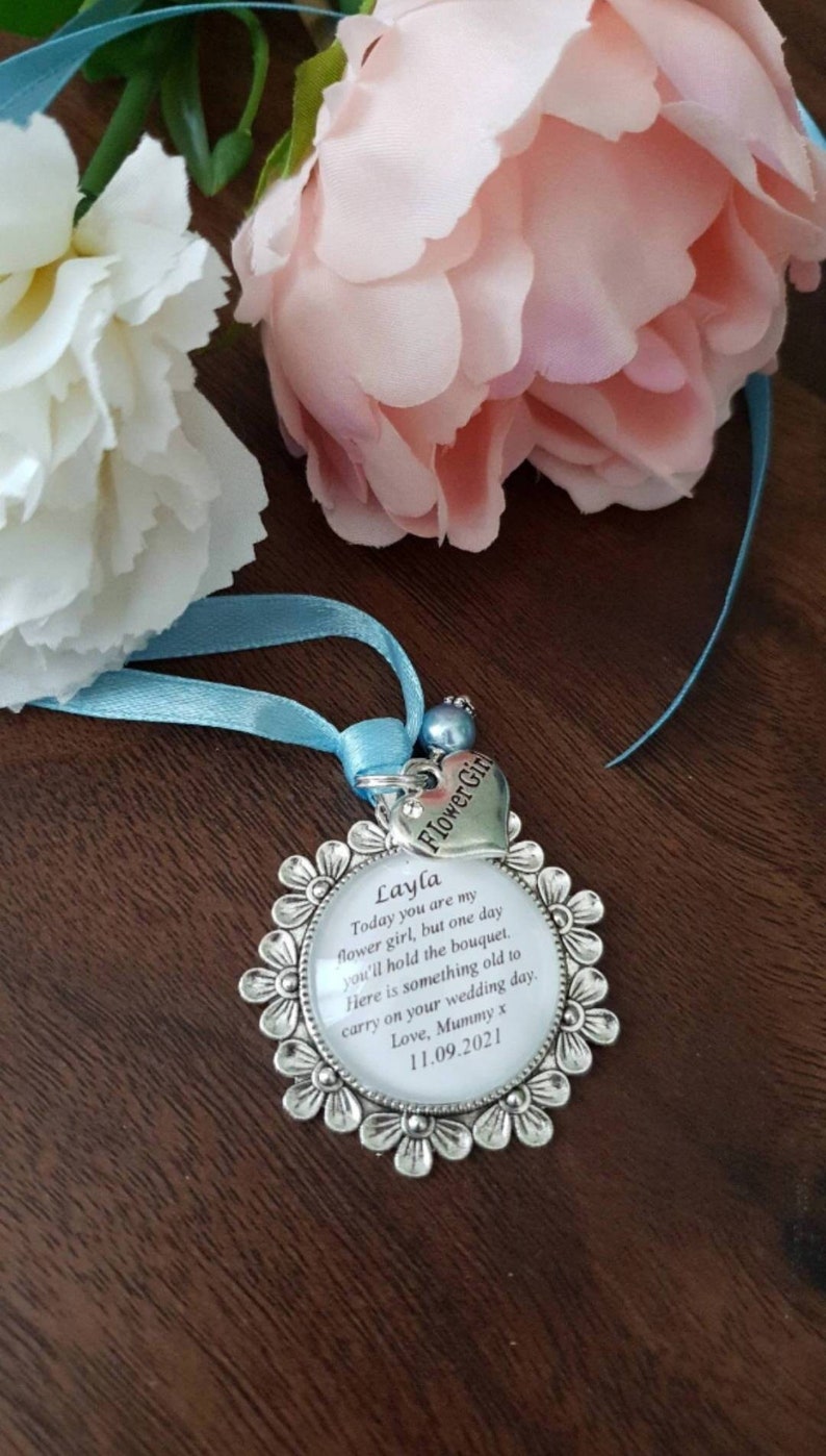 K&ouml;nnte beinhalten: Ein silberner Anh&auml;nger mit wei&szlig;em Hintergrund und schwarzem Text, der lautet: "Layla Heute bist du meine Blumenm&auml;dchen, aber eines Tages wirst du den Strau&szlig; halten. Hier ist etwas Altes, das du an deinem Hochzeitstag tragen kannst. Liebe, Mama x 11.09.2021". Der Anh&auml;nger ist an einem blauen Band und einem silbernen Herzanh&auml;nger befestigt, auf dem "Blumenm&auml;dchen" steht.