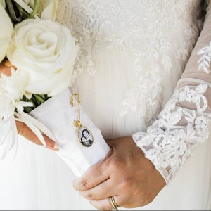Könnte beinhalten: Ein weißer Brautstrauß mit weißen Rosen und weißen Blumen, gehalten von einer Braut in einem weißen Spitzenkleid. Eine goldene Sicherheitsnadel mit einem kleinen Foto-Anhänger ist am Strauß befestigt.