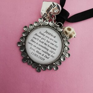 May include: A silver charm with a white background and black text that reads "Jessica Today you are our flower girl, but one day you'll hold the bouquet. Here is something old to carry on your wedding day. Love, Mum & Dad 11/05/2020". The charm is attached to a black ribbon and a silver heart charm that reads "Flower Girl".