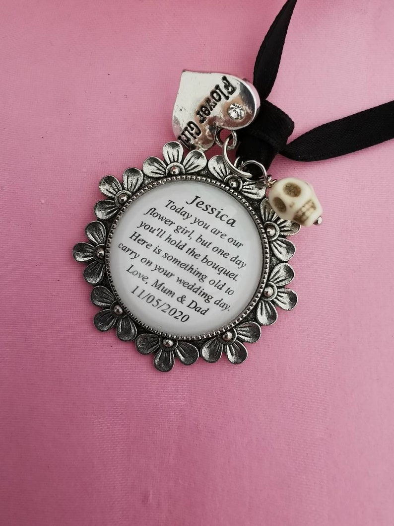 May include: A silver charm with a white background and black text that reads "Jessica Today you are our flower girl, but one day you'll hold the bouquet. Here is something old to carry on your wedding day. Love, Mum & Dad 11/05/2020". The charm is attached to a black ribbon and a silver heart charm that reads "Flower Girl".