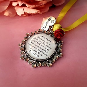 May include: A silver charm with a white background and a message that reads "Today you are our flower girl. But one day you'll hold the bouquet, here is something old to carry on your wedding day. Love, Aunt Kayla 21.06.2019". The charm is surrounded by a silver frame with flower designs. The charm is attached to a yellow ribbon with a red flower.