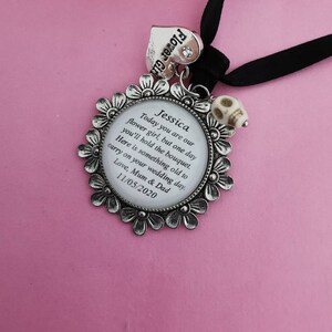 May include: A silver flower girl charm with a white enamel circle containing the text "Jessica Today you are our flower girl, but one day you'll hold the bouquet. Here is something old to carry on your wedding day. Love, Mum & Dad 11/05/2020". The charm is attached to a black ribbon with a silver heart charm that says "Flower Girl".