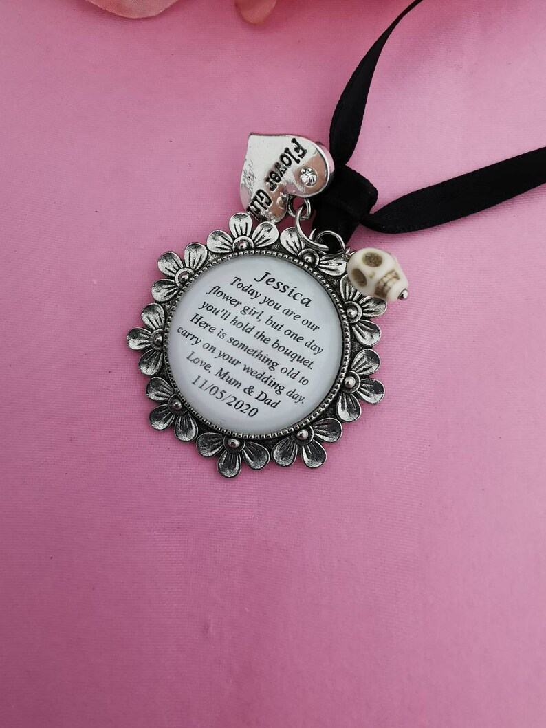 May include: A silver flower girl charm with a white enamel circle containing the text "Jessica Today you are our flower girl, but one day you'll hold the bouquet. Here is something old to carry on your wedding day. Love, Mum & Dad 11/05/2020". The charm is attached to a black ribbon with a silver heart charm that says "Flower Girl".