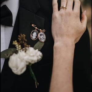May include: A groom wearing a black tuxedo jacket with a white bow tie and a boutonniere. The boutonniere features two small oval-shaped photo charms with gold frames. The bride's hand is resting on the groom's shoulder, and she is wearing a gold ring with a large diamond.