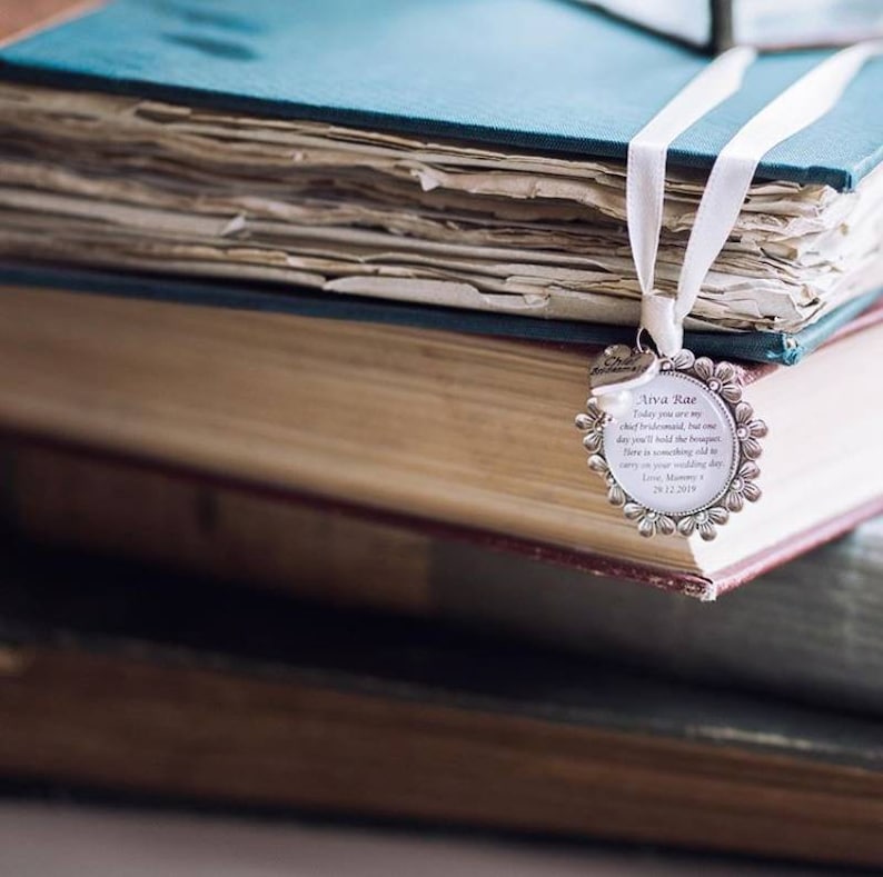 K&ouml;nnte beinhalten: Ein silbernes Lesezeichen mit floralem Design und einer personalisierten Botschaft f&uuml;r eine Brautjungfer. Das Lesezeichen h&auml;ngt an einem Stapel alter B&uuml;cher.