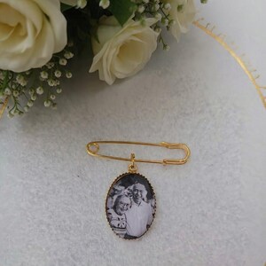 Może przedstawiać: Złota agrafka z owalnym wisiorkiem z czarno-białym zdjęciem pary. Szpilka jest umieszczona na białej powierzchni, z białymi różami w tle.