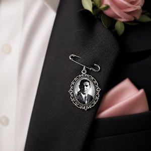 May include: A silver memorial pin with a black and white portrait of a man, attached to a black suit jacket with a safety pin. A pink pocket square and a pink rose are also visible.