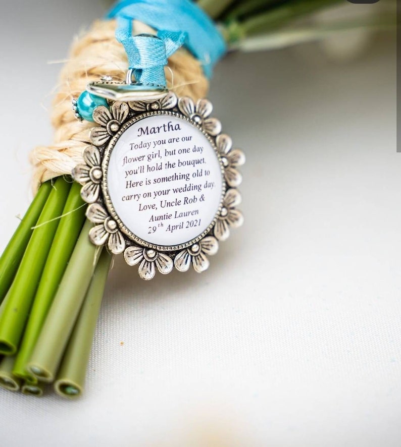 K&ouml;nnte beinhalten: Ein silberner Anh&auml;nger mit wei&szlig;em Hintergrund und schwarzem Text, der "Martha Today you are our flower girl, but one day you'll hold the bouquet. Here is something old to carry on your wedding day Love, Uncle Rob & Auntie Lauren 29th April 2021" lautet.