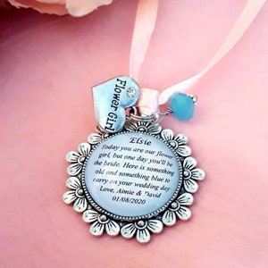 May include: A silver charm with a white background and a message for a flower girl. The charm reads "Elsie Today you are our flower girl, but one day you'll be the bride. Here is something old and something blue to carry on your wedding day Love, Aimie & David 01/08/2020".