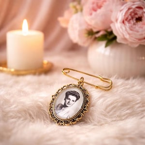 May include: A gold brooch with an oval pendant displaying a black and white portrait of a woman. The brooch has an ornate, filigree frame and a safety pin fastening. A lit candle and a vase of pink flowers are in the background.