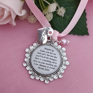 K&ouml;nnte beinhalten: Ein silberner Anh&auml;nger mit wei&szlig;em Hintergrund und schwarzem Text, der "Nancy Today you are our baby girl, but one day you'll hold the bouquet. Here is something old to carry on your wedding day. Love, Mummy & Daddy 09.08.2024" lautet. Der Anh&auml;nger ist an einem rosa Band mit einem kleinen rosa Herzanh&auml;nger befestigt, auf dem "Baby Girl" steht.