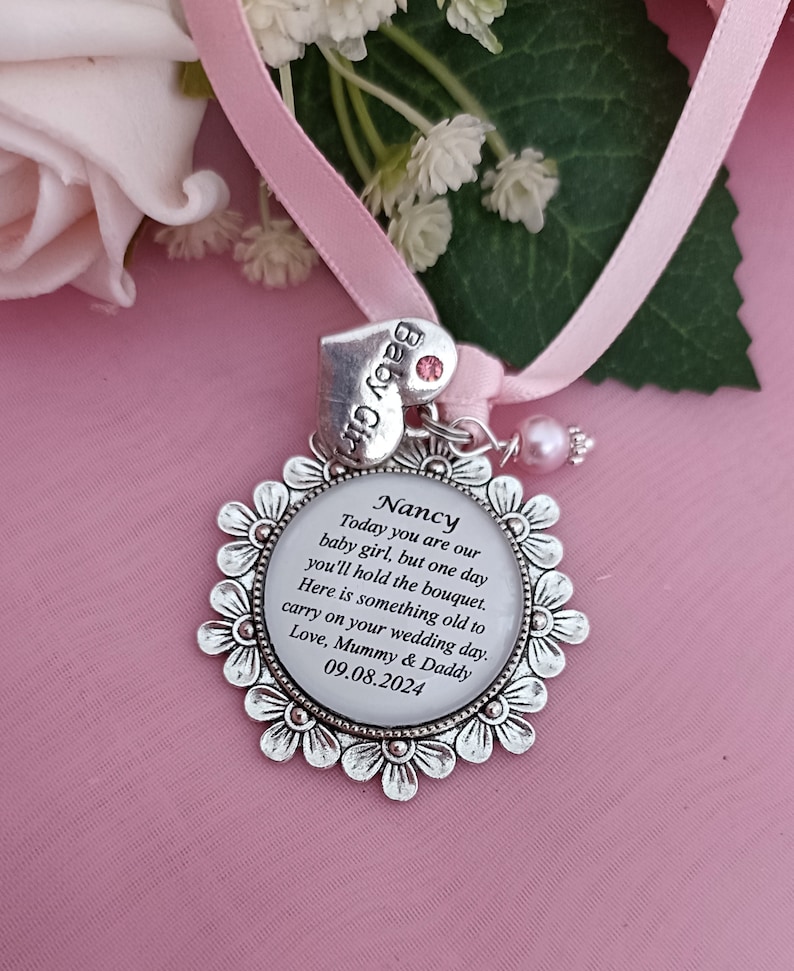 K&ouml;nnte beinhalten: Ein silberner Anh&auml;nger mit wei&szlig;em Hintergrund und schwarzem Text, der "Nancy Today you are our baby girl, but one day you'll hold the bouquet. Here is something old to carry on your wedding day. Love, Mummy & Daddy 09.08.2024" lautet. Der Anh&auml;nger ist an einem rosa Band mit einem kleinen rosa Herzanh&auml;nger befestigt, auf dem "Baby Girl" steht.