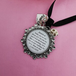 May include: A silver charm with a white background and black text. The charm features a floral design and the text "Jessica Today you are our flower girl, but one day you'll hold the bouquet. Here is something old to carry on your wedding day. Love, Mum & Dad 11/05/2020".