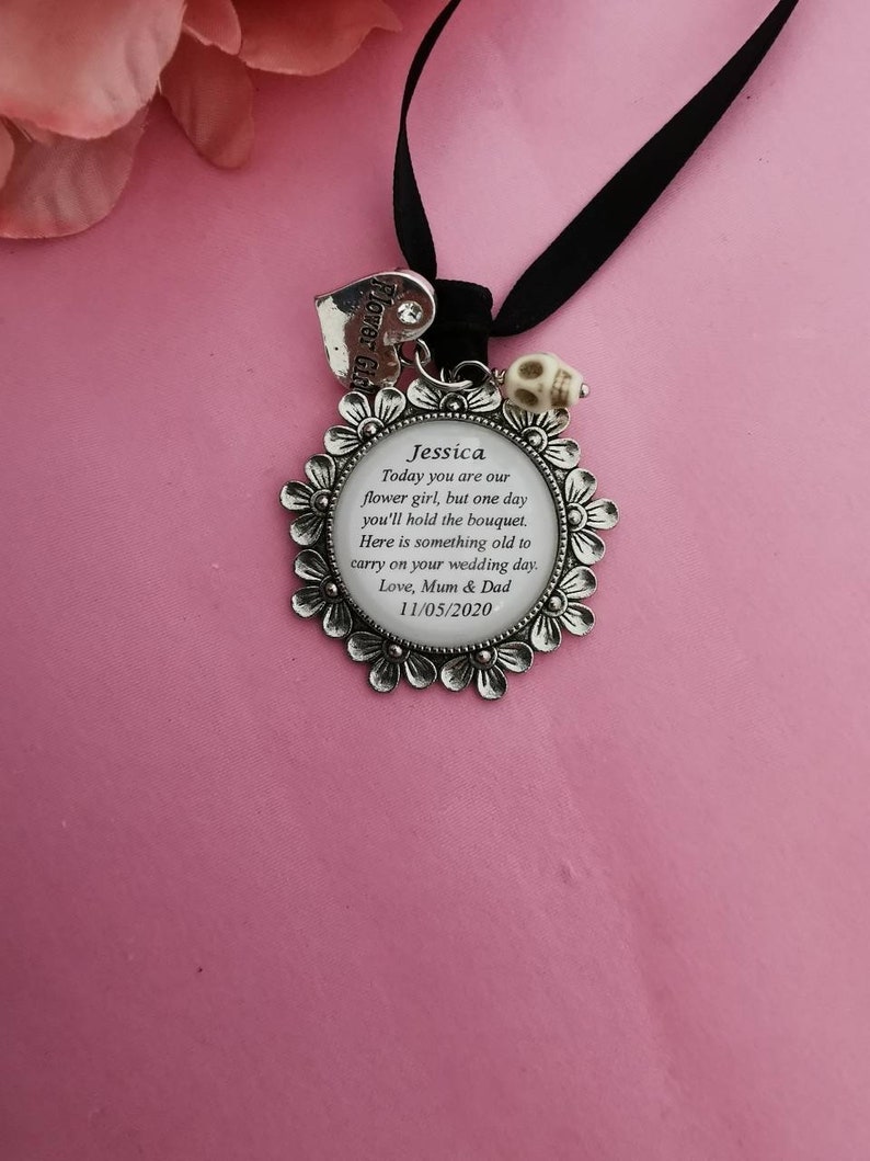 May include: Silver charm with a floral design and a white skull bead. The charm has a clear glass center with the text "Jessica Today you are our flower girl, but one day you'll hold the bouquet. Here is something old to carry on your wedding day. Love, Mum & Dad 11/05/2020".