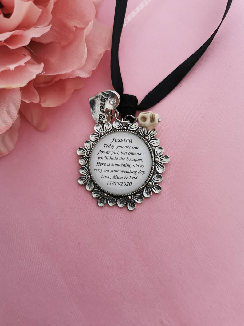 May include: A silver charm with a floral design and a message for a flower girl. The charm reads "Jessica Today you are our flower girl, but one day you'll hold the bouquet. Here is something old to carry on your wedding day. Love, Mum & Dad 11/05/2020".