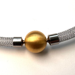 Op de afbeelding: Een zilveren mesh ketting met een goudkleurige bol in het midden. De ketting heeft zilverkleurige metalen accenten aan weerszijden van de bol. De ketting wordt gepresenteerd tegen een witte achtergrond.
