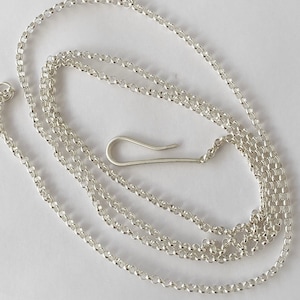 Könnte beinhalten: Eine silberfarbene Halskette mit einer filigranen Kette und einem Hakenverschluss. Die Kette besteht aus kleinen, runden Gliedern. Der Hakenverschluss hat ein einfaches, gebogenes Design. Die Halskette wird vor weißem Hintergrund präsentiert.