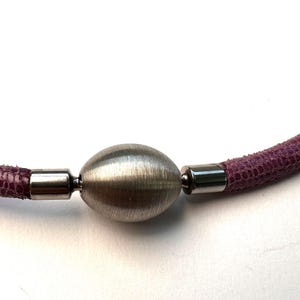 Op de afbeelding: Een close-up van een ketting met een getextureerd, paars koord en een ovale hanger van geborsteld zilver. Het koord is met zilveren metalen sluitingen aan de hanger bevestigd. De ketting staat tegen een witte achtergrond.