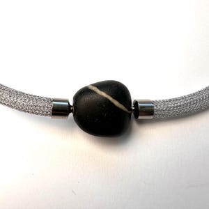 Op de afbeelding: Zilveren mesh ketting met een zwarte stenen hanger. De steen heeft een witte streep en wordt op zijn plaats gehouden door zilveren metalen sluitingen. Op een witte achtergrond.