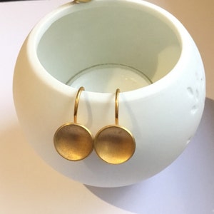 Pode incluir: Um par de brincos dourados com um formato redondo ligeiramente côncavo. Os brincos estão pendurados em um simples laço de arame.