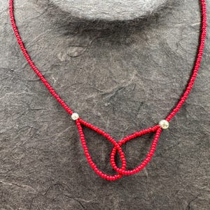 Könnte beinhalten: Eine rote Perlenkette mit einer silbernen Perle an jedem Ende. Die Kette ist in der Mitte zu einem Knoten gebunden, der eine Schmetterlingsform bildet.