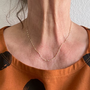 May include: A delicate gold chain necklace with a fine, beaded design. The necklace is worn by a person with a light brown shirt.