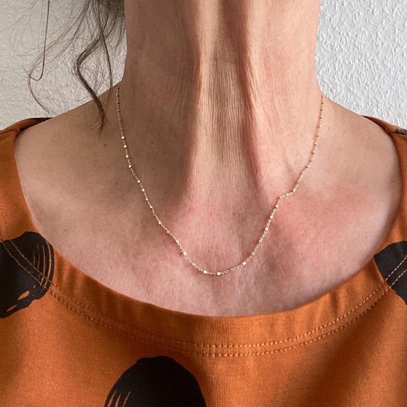 May include: A delicate gold chain necklace with a fine, beaded design. The necklace is worn by a person with a light brown shirt.