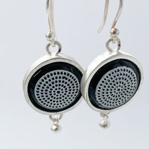 Può includere: Un paio di orecchini d'argento con disegni circolari in bianco e nero. Ogni orecchino ha una piccola sfera d'argento che pende dalla parte inferiore.